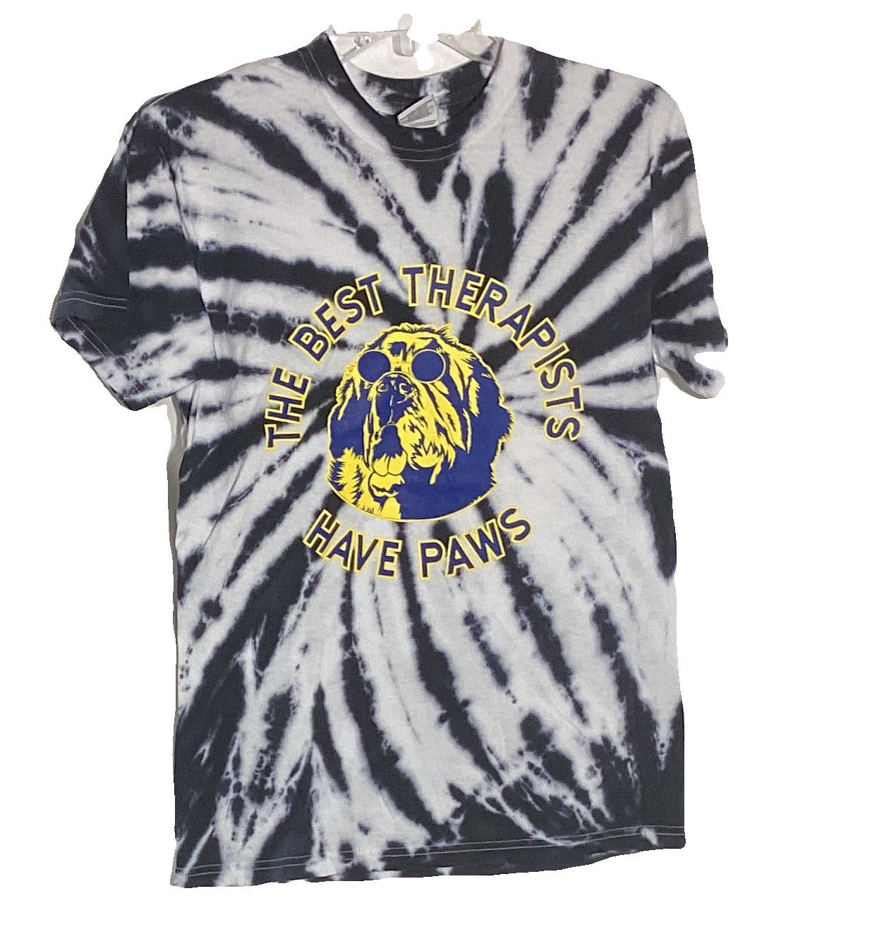 Alt text: T-shirt with black and white tie-dye design featuring a graphic print saying "The Best Therapists Have Paws" with an illustration of a golden retriever wearing sunglasses, framed by a circular yellow border.