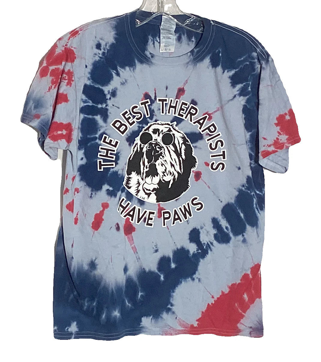 Alt text: "4th of July themed tie-dye t-shirt with a graphic saying 'The Best Therapists Have Paws' featuring an illustration of a dog wearing sunglasses in red, white, and blue colors, displayed on a hanger
